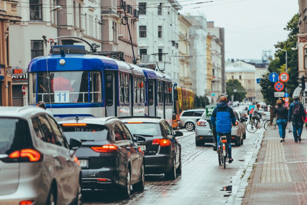 Modern vehicles driving slowly with riding cyclist on busy road in city while people walking and sidewalk near old buildings